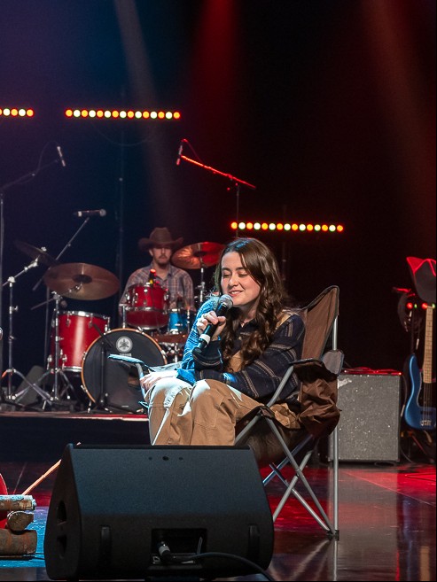 Maelie Berthelet assise dans une chaise de camping sur grande scène, micro à la main